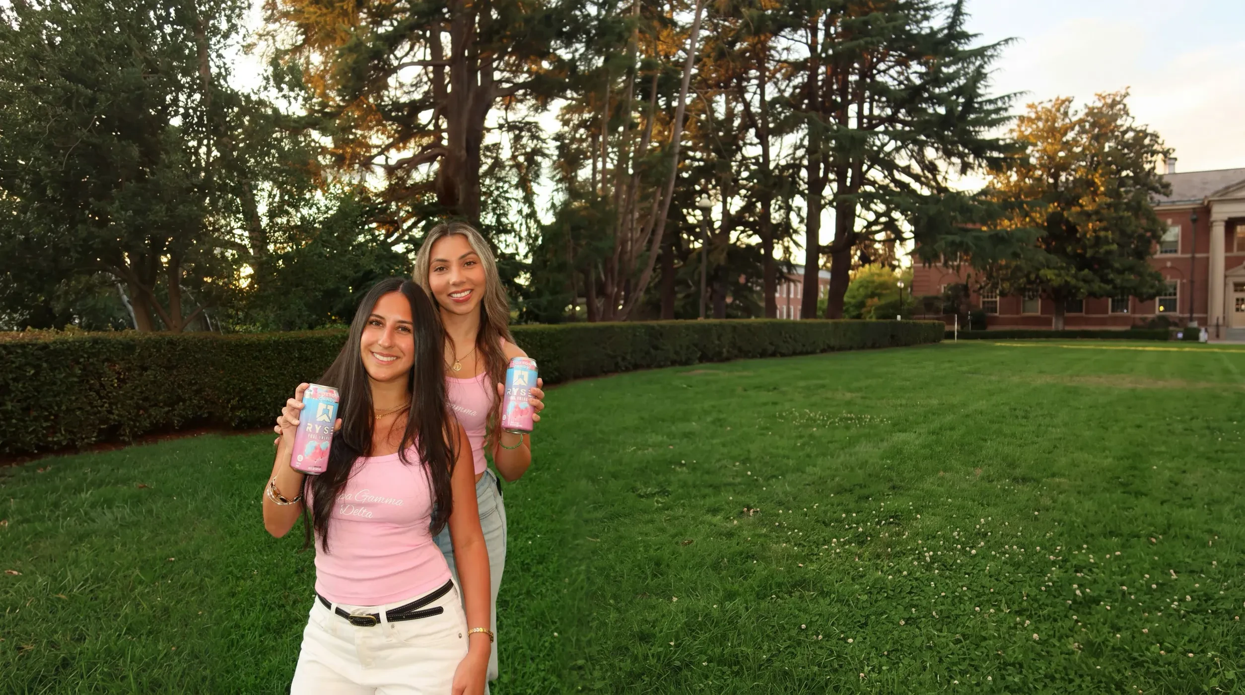 Two sorority sisters in pink tops holding cans of RYSE on a campus lawn at golden hour