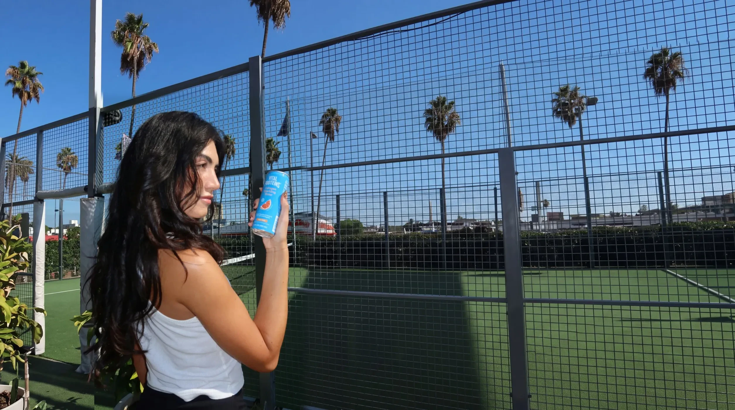 A woman holding a can of Vital Proteins at a sunny outdoor padel court lined with palms