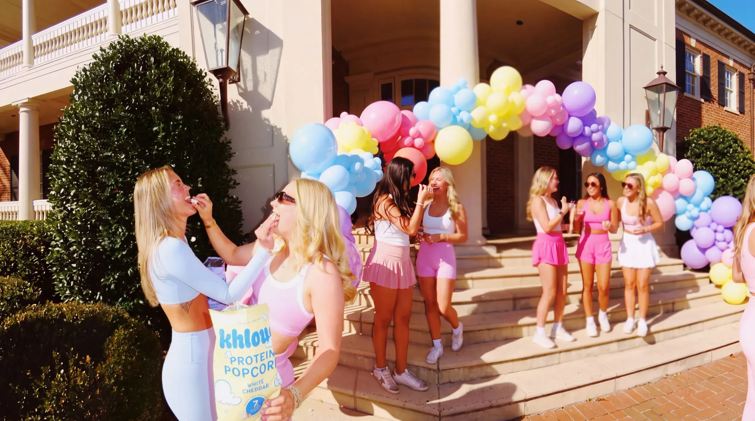 Two sorority sisters feeding each other Khloud protein popcorn under a balloon arch on the house steps